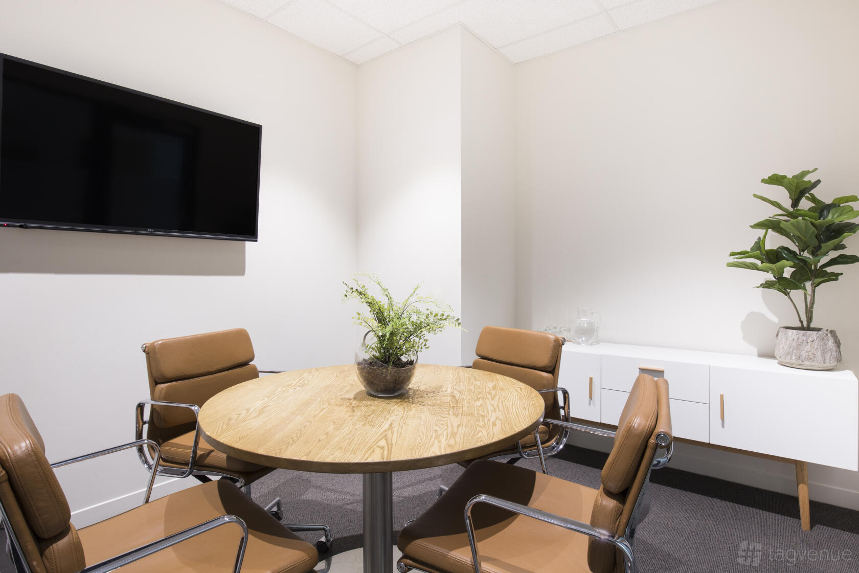 A meeting room with a round wooden table, four leather chairs, wall-mounted TV, and sideboard with plants at Corporate One.