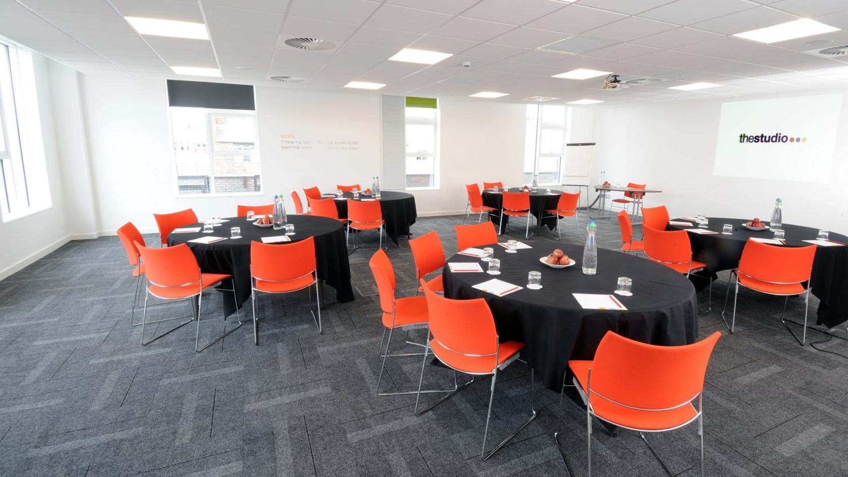 A conference centre with round tables, bright orange chairs, and large windows at The Studio Manchester.