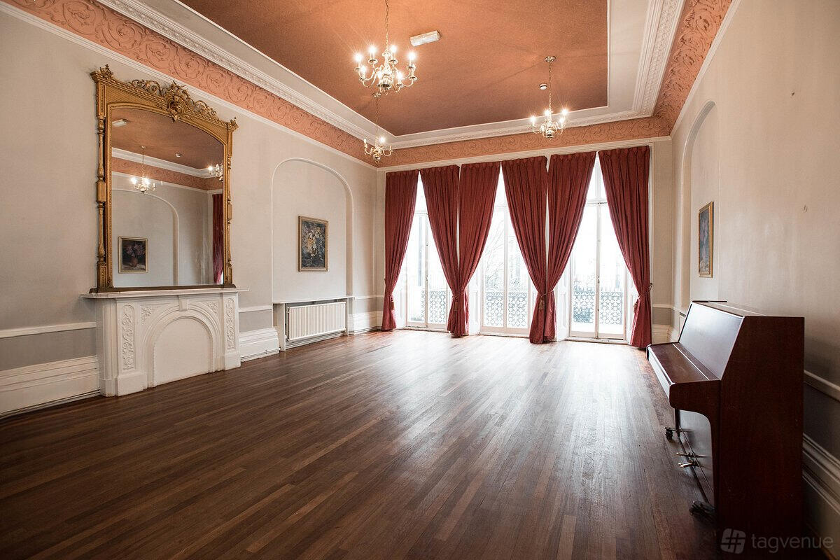 A function room with tall windows, red drapes, a grand mirror above a white fireplace, and a piano at 72 QT Guest House London.