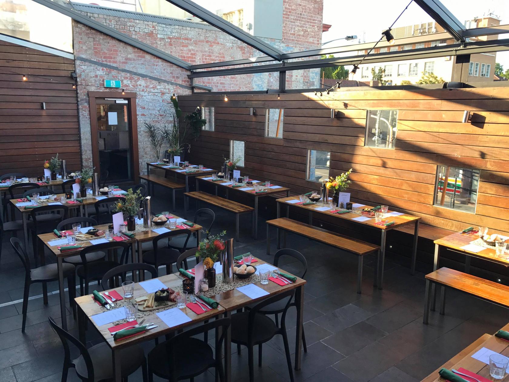A rooftop bar with wooden walls, glass ceiling, and long tables set for dining at Arcadia Hotel.