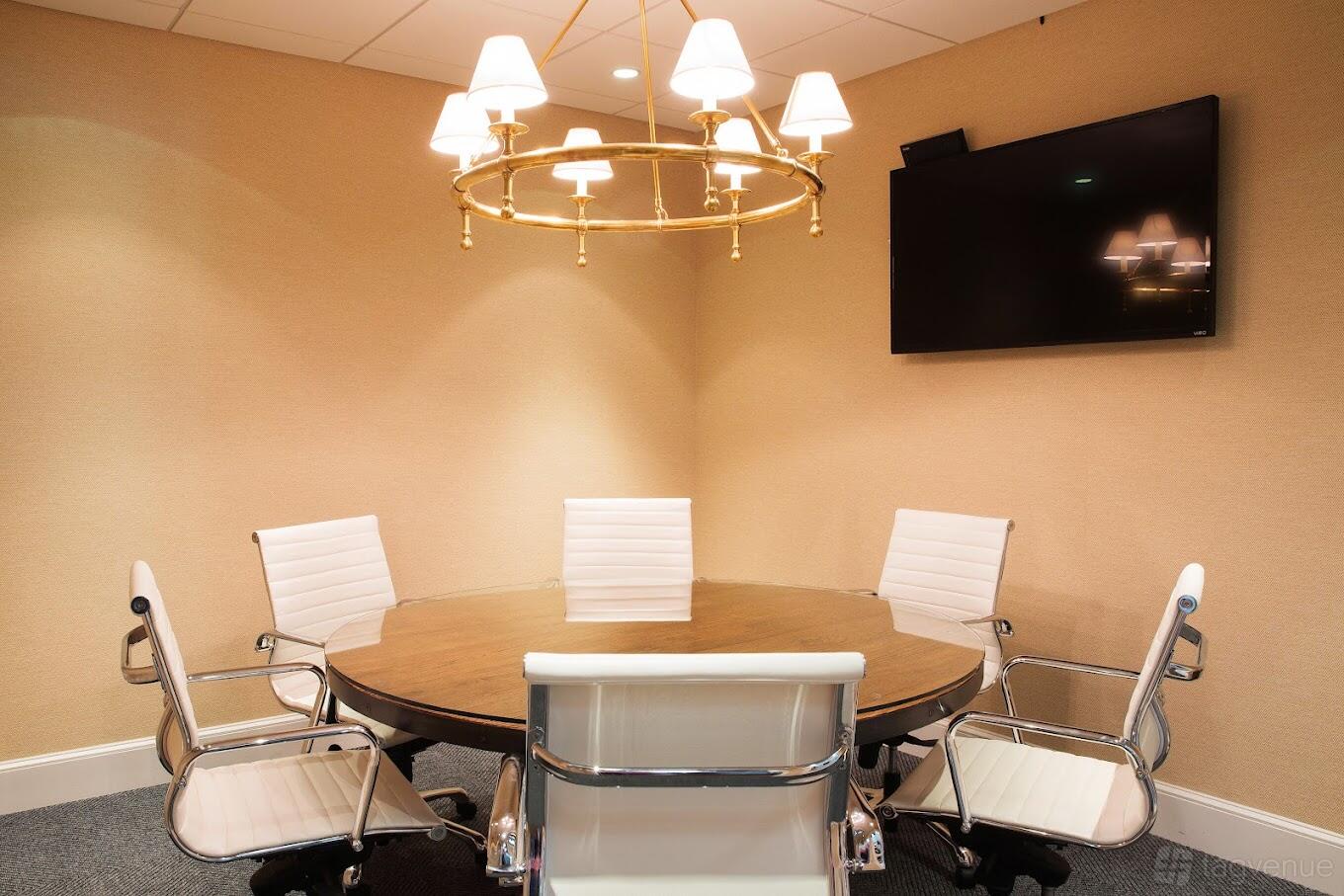 A meeting room with a round wooden table, white chairs, wall-mounted TV, and brass chandelier at Work Station Cohasset.