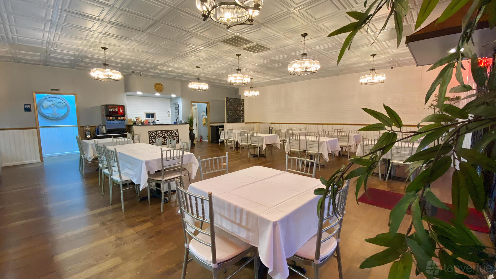 A dining room with white tablecloths, wooden floors, and silver chairs under chandelier lighting at Lazeeza Grill Restaurant & Banquet Hall.