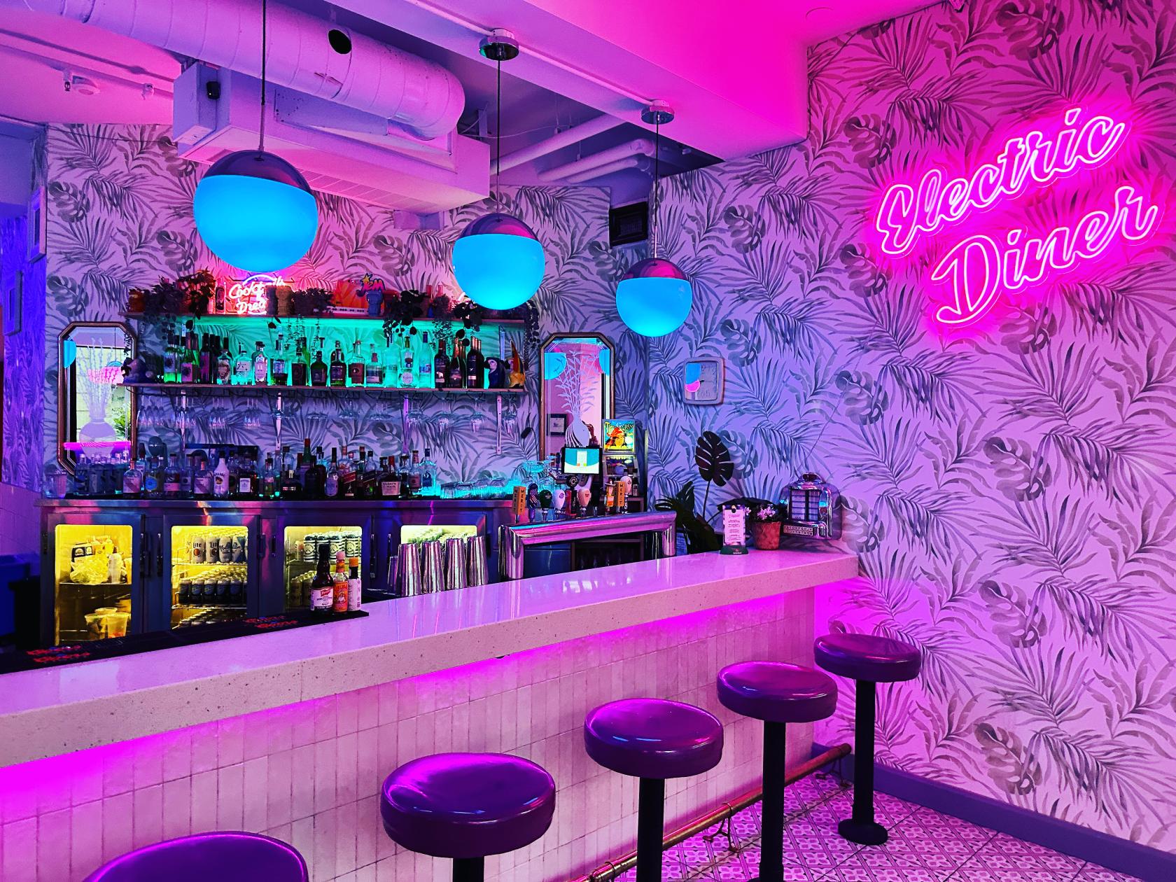 A restaurant bar with neon lighting, patterned wallpaper, globe pendant lights, and a pink neon sign at Electric Diner.