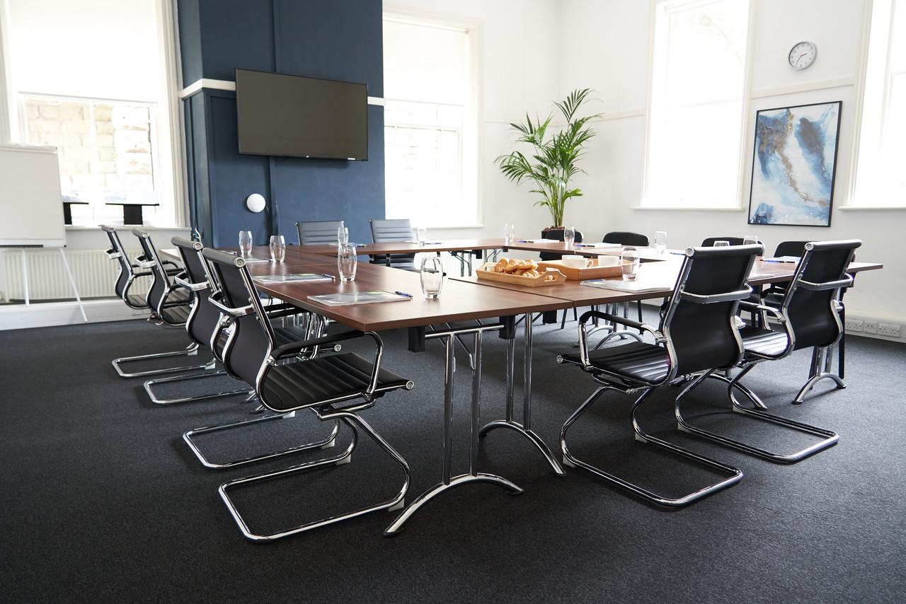 A training room with U-shaped tables, black chairs, a wall-mounted screen, and large windows at Clavering House.
