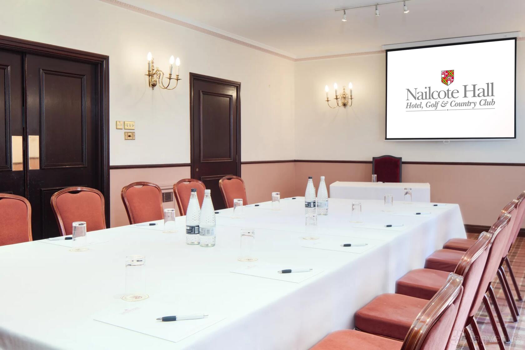 An event space with a U-shaped table setup, cushioned chairs, and a projector screen at Nailcote Hall Hotel.