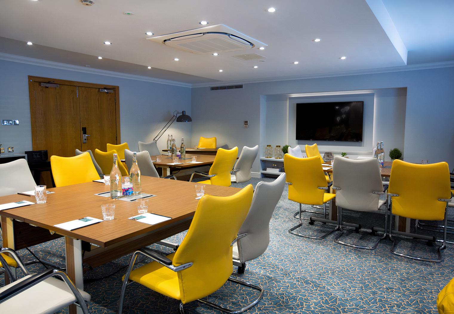 A hotel meeting room with yellow and white chairs, wooden tables, and a wall-mounted screen at Aztec Hotel & Spa.