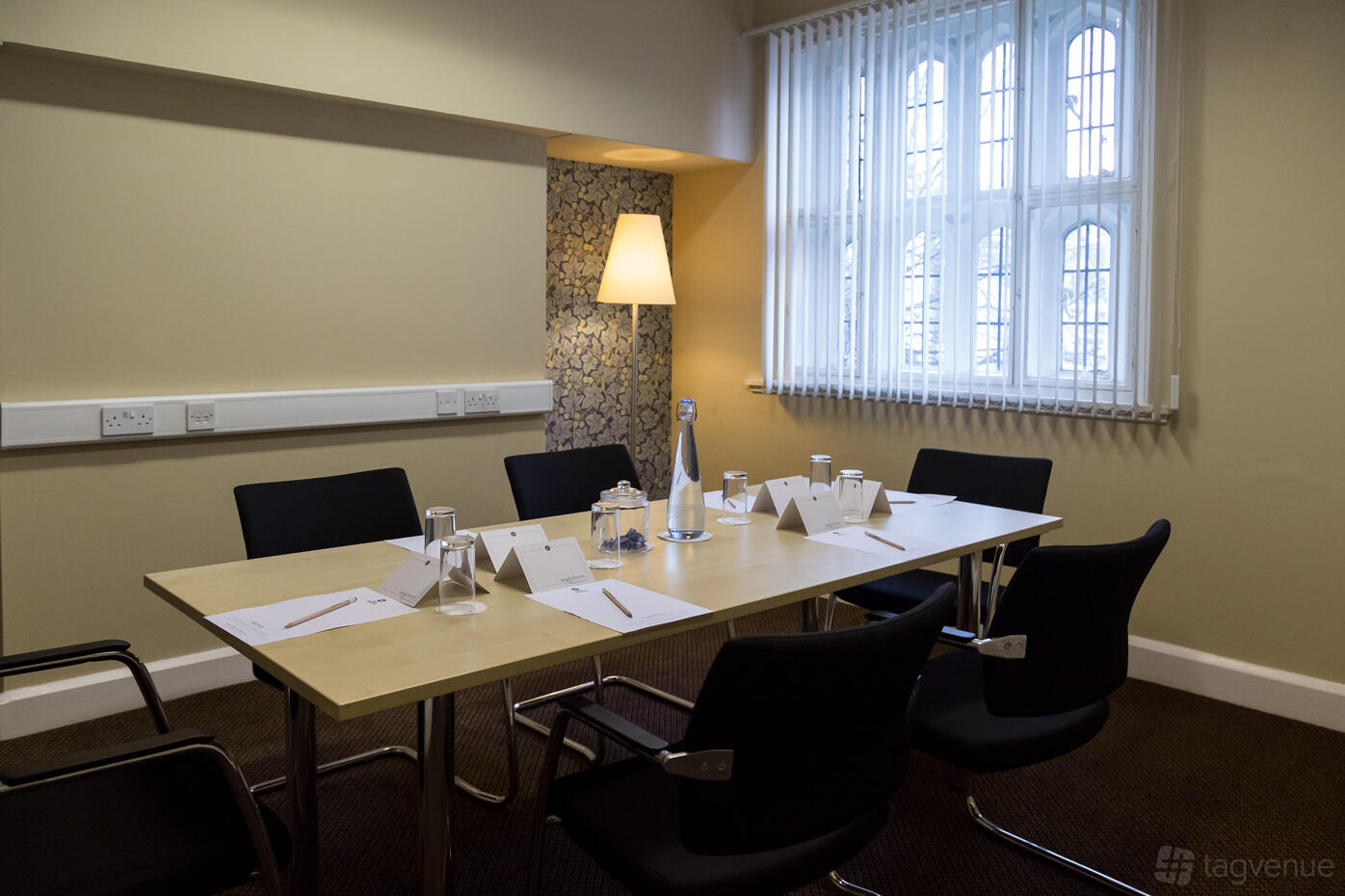 A meeting room with a rectangular table, black chairs, bottled water, and natural light from arched windows at Plough & Harrow Hotel.