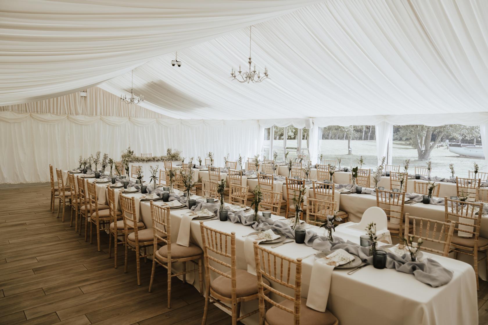 A marquee hall with draped white ceilings, chandeliers, and banquet tables with natural wood chairs at Crown Hall Farm.