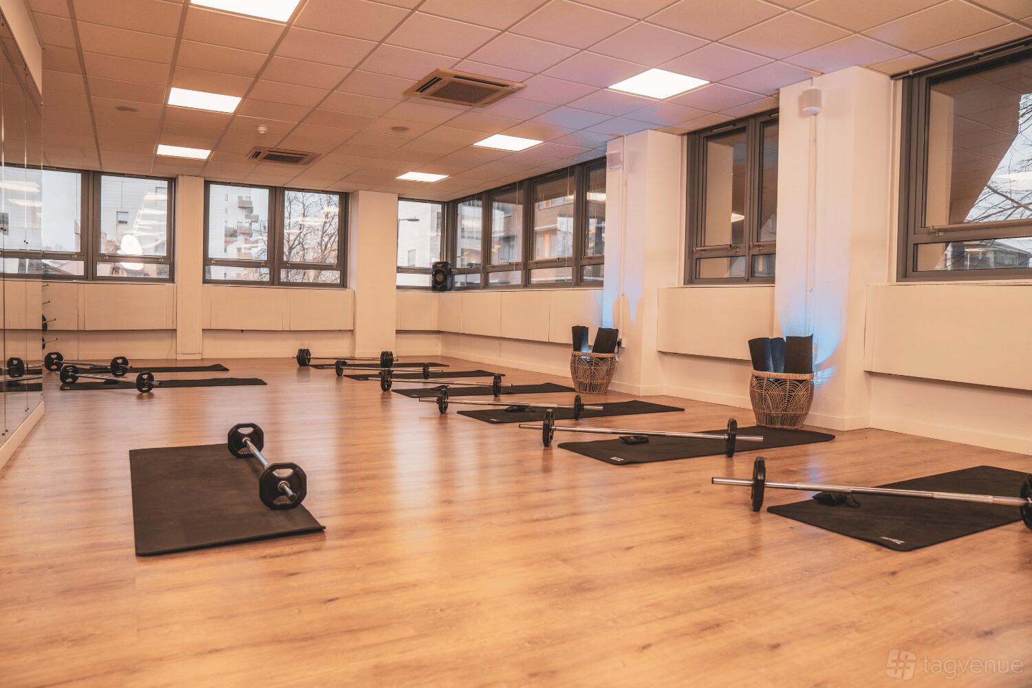 A fitness studio with wood flooring, floor-to-ceiling windows, mirrors, and workout mats with barbells at MAKE IT Barking.