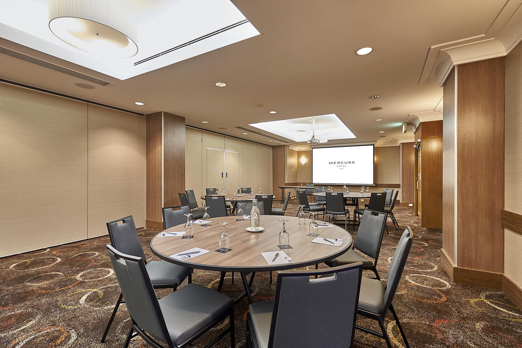 A function room with round tables, cushioned chairs, and built-in projector screen at Mercure Perth.