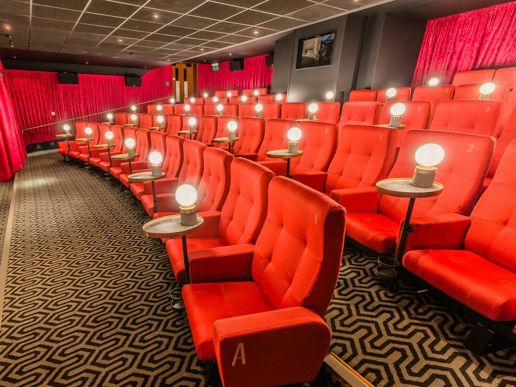 A cinema with red plush seats, small round side tables, and patterned carpet at Curzon Mayfair.