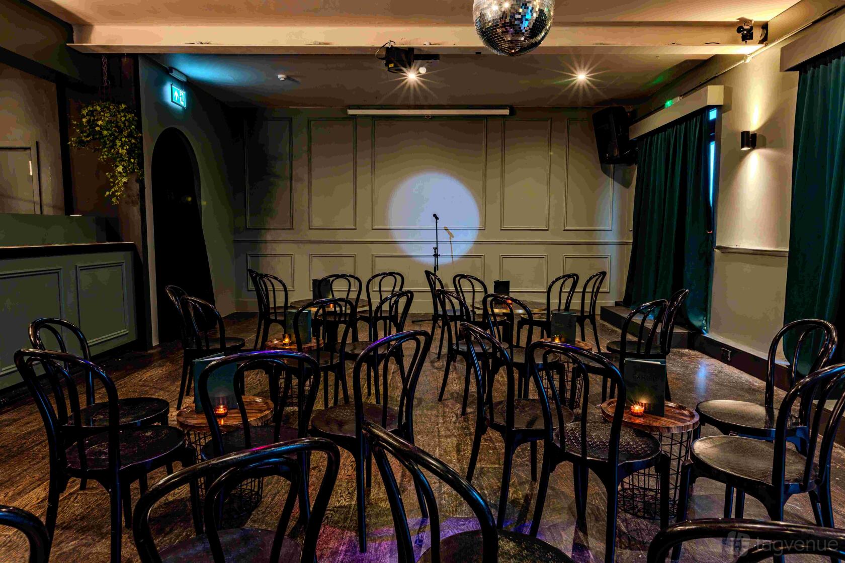 An event space with rows of black chairs, a spotlighted stage, dark curtains, and a disco ball at Exhibit.