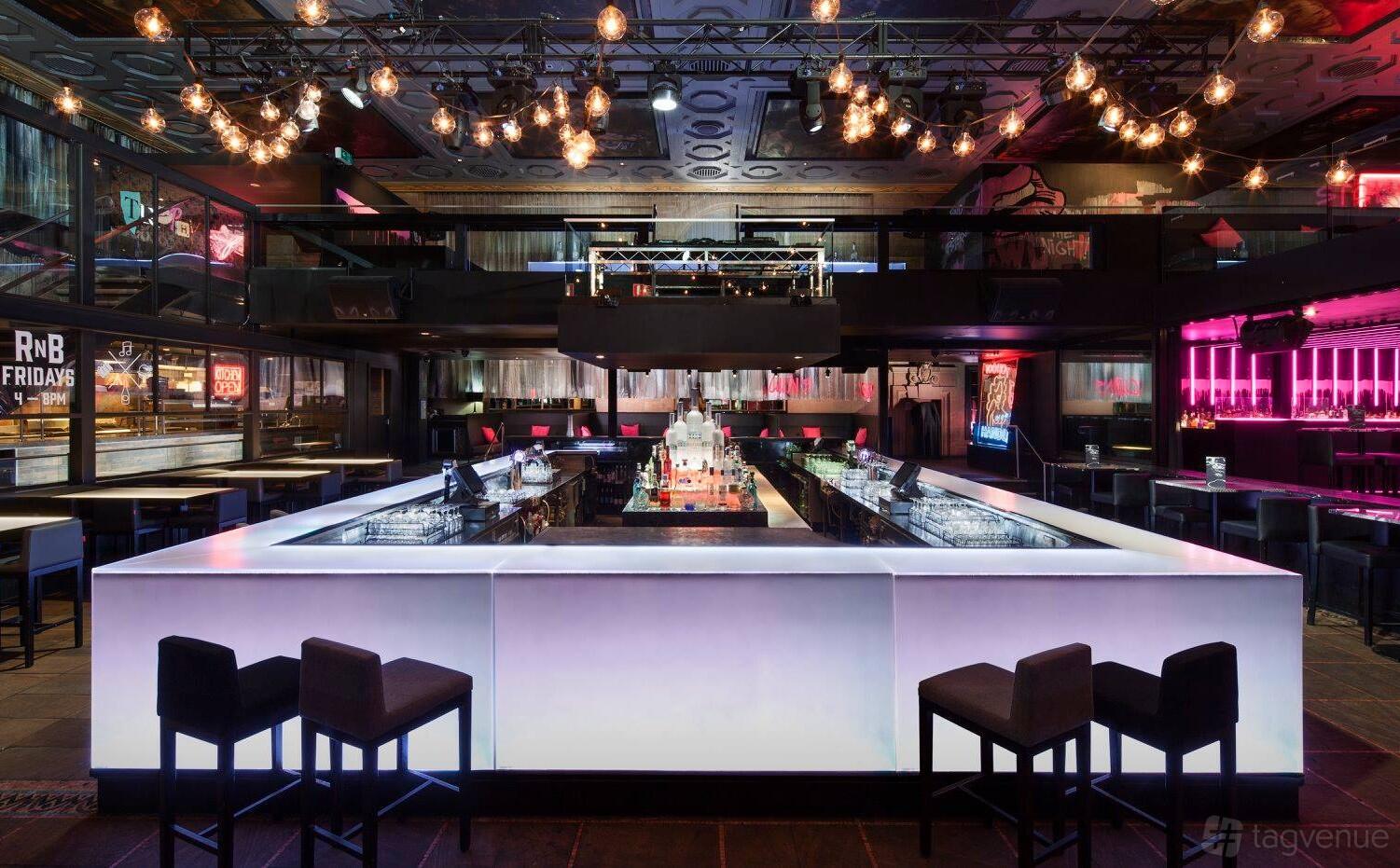 A nightclub with an illuminated square bar, modern seating, and overhead string lights at Ms Collins.