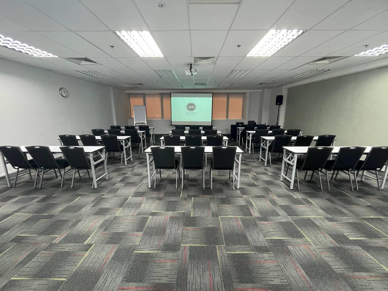 An auditorium with rows of black chairs, white tables, and a projector screen at RNN Conference Centre.