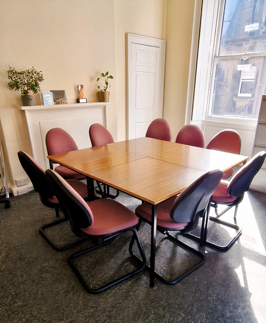 A meeting room with a square wooden table, eight pink chairs, and natural light from a bay window at Birthlink.