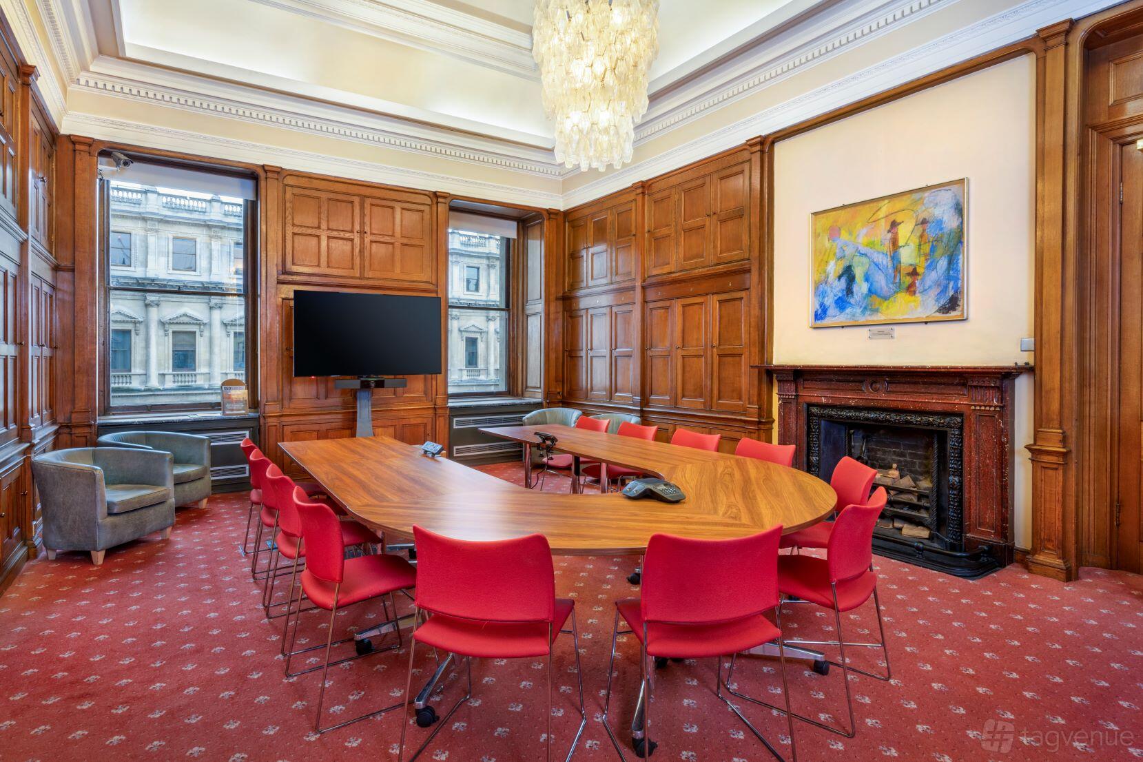 A meeting room with a large oval table, red chairs, wood paneling, fireplace, and chandelier at The Geological Society.