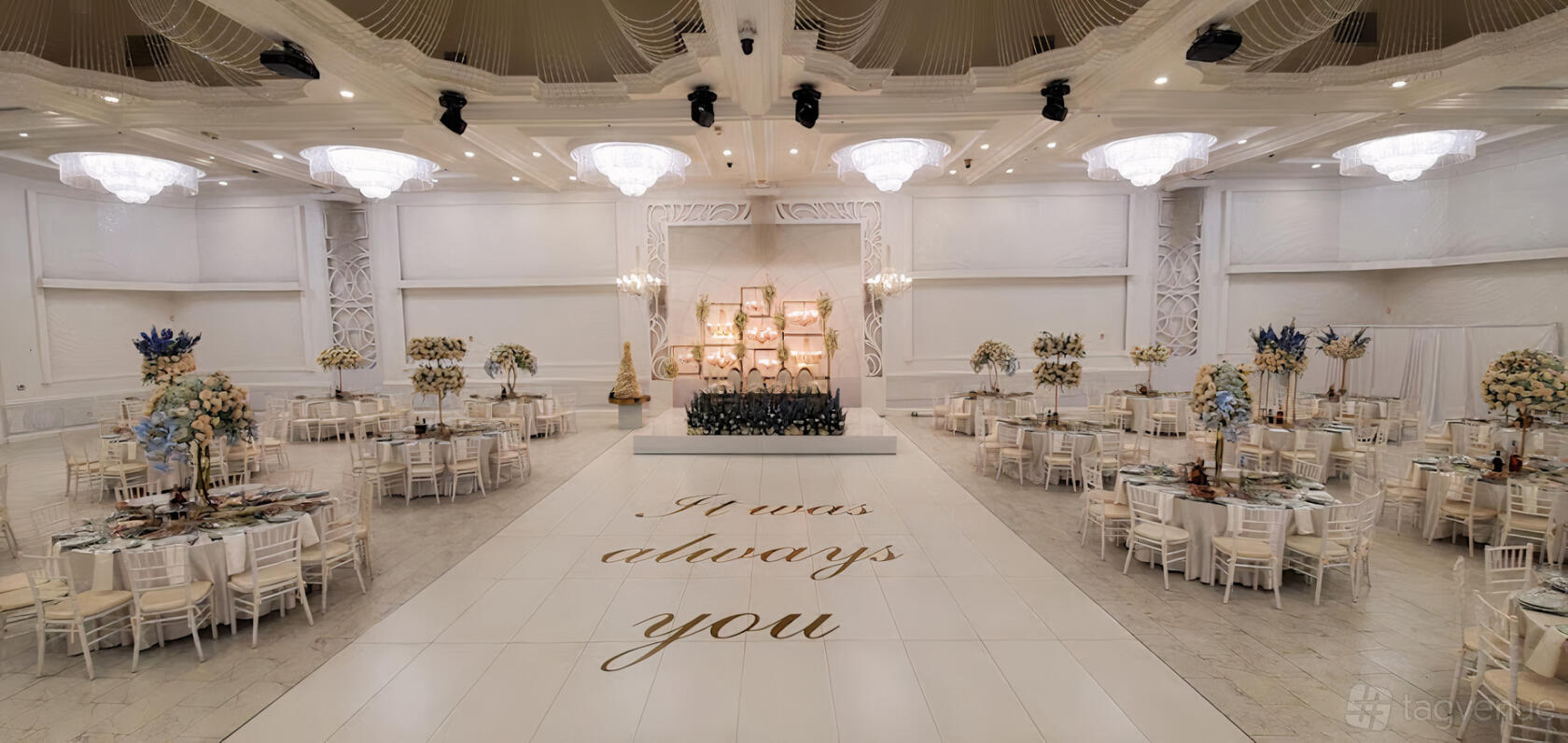 A banquet hall with crystal chandeliers, white walls, round tables with floral centerpieces, and a dance floor at The Great Caesar Banquet Hall.