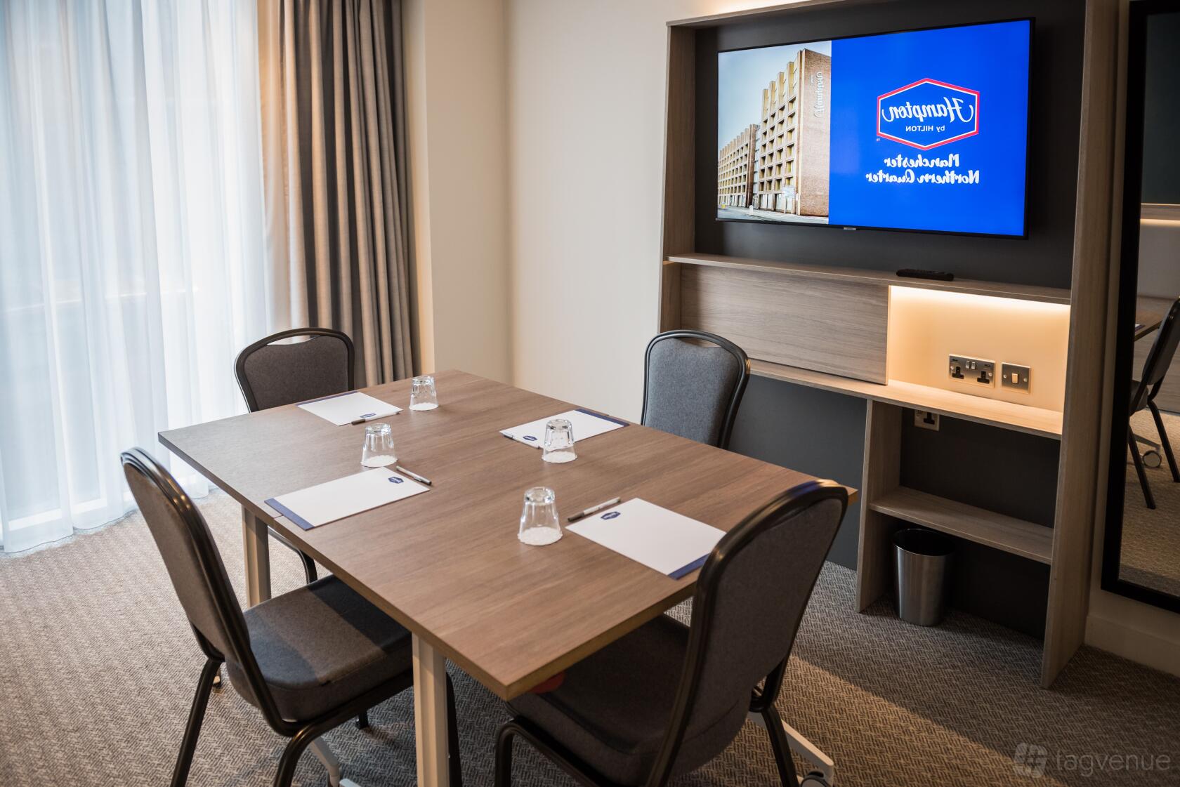 A meeting room with a wooden table, four chairs, notepads, glasses, and a wall-mounted screen at Hampton by Hilton Manchester Northern Quarter.