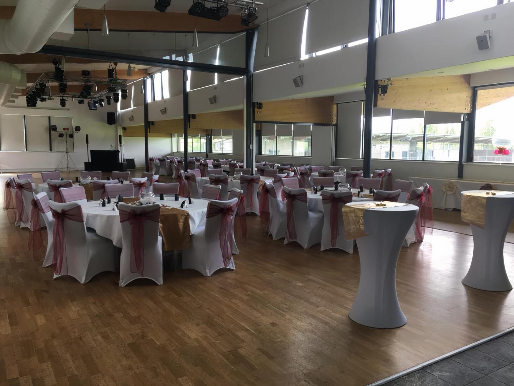 A function room with round tables draped in white linens and red sashes, wooden floors, and tall windows at Princes Park.