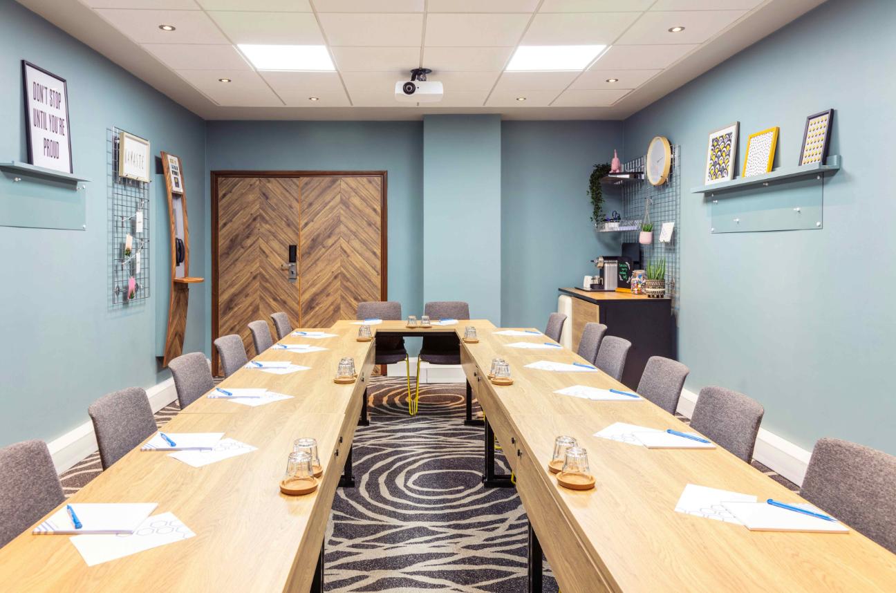 A meeting room with U-shaped wooden tables, gray chairs, wall decor, and a beverage station at Novotel Edinburgh Centre.