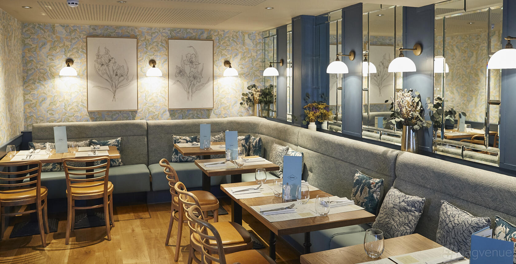 A function room with upholstered booth seating, wooden chairs, floral wallpaper, and wall-mounted lamps at Cote Brasserie St. Christopher's Place.