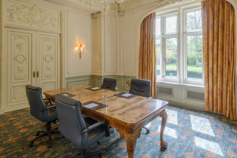 A hotel meeting room with ornate wall moldings, a carved wood table, and large windows with gold curtains at Warren House Hotel.