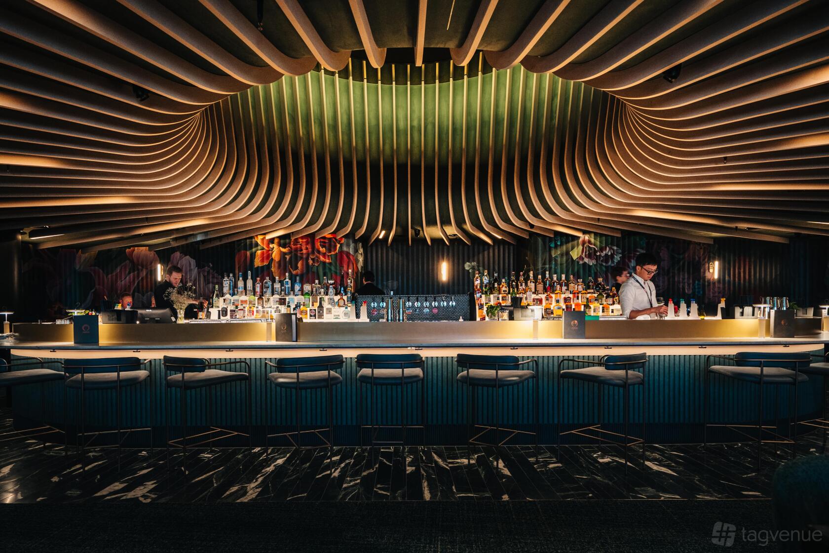 A restaurant bar with sculptural wooden ceiling panels, backlit shelves of liquor, and modern bar stools at Amphora.