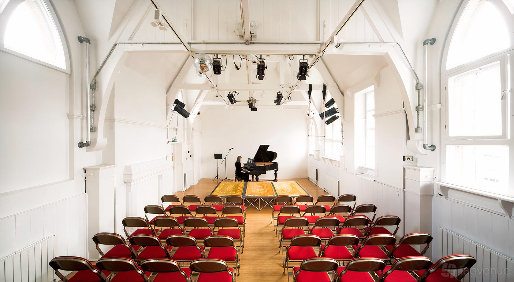 A gallery hall with a raised stage, grand piano, arched windows, and rows of red chairs at October Gallery.