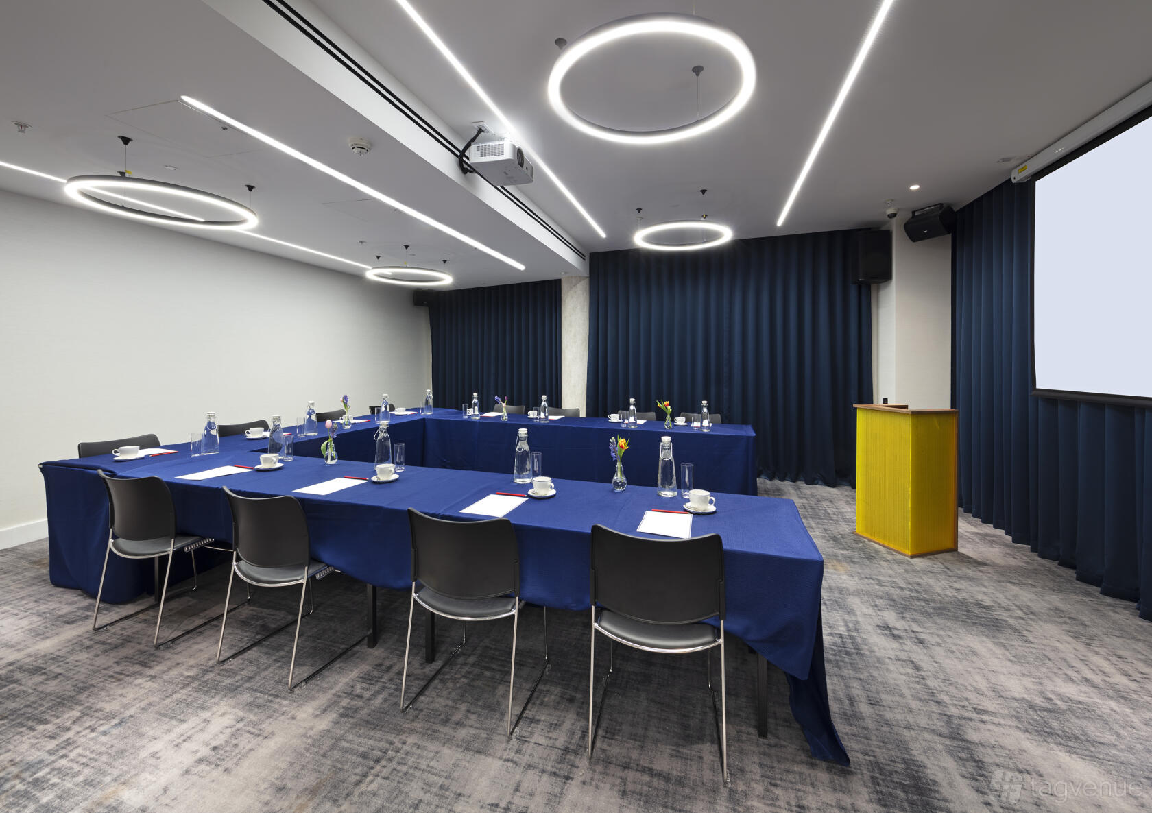 An event space in a hotel with ring ceiling lights, navy blue tablecloths, and a yellow podium at Tower Suites by Blue Orchid.