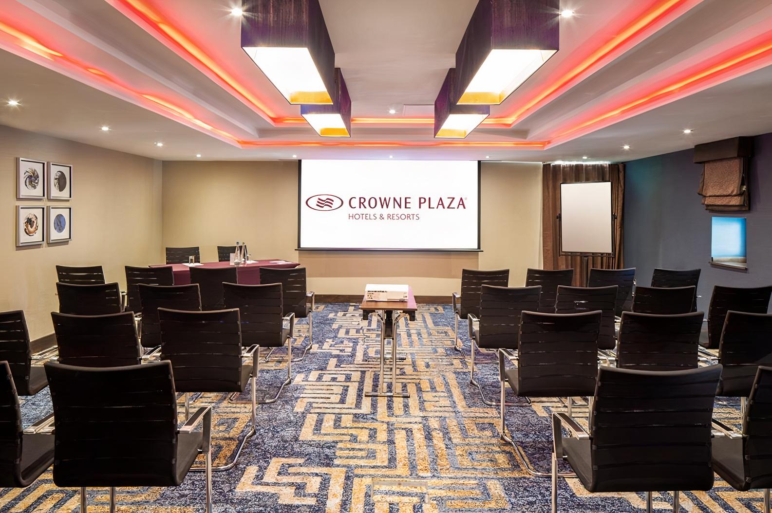 A hotel meeting room with rows of black chairs, patterned carpet, and ceiling lights at Crowne Plaza London Gatwick.