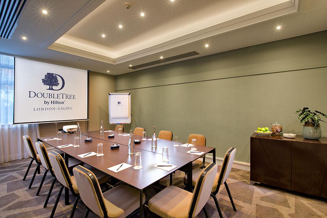 A hotel boardroom with a square meeting table, ergonomic chairs, projection screen, and natural lighting at DoubleTree by Hilton London - Ealing.