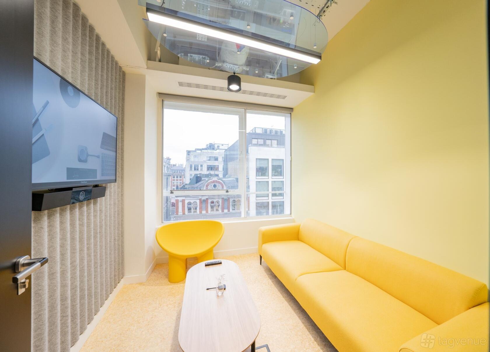 A meeting room with yellow seating, a large window, flat-screen display, and acoustic wall panels at Huckletree Oxford Circus.