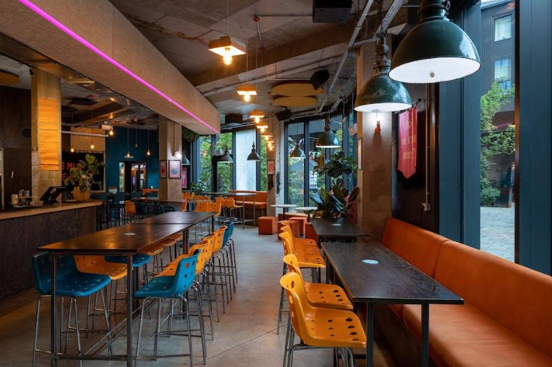 A restaurant with long wooden tables, yellow chairs, exposed ceiling, and large windows at Nell's at Kampus.