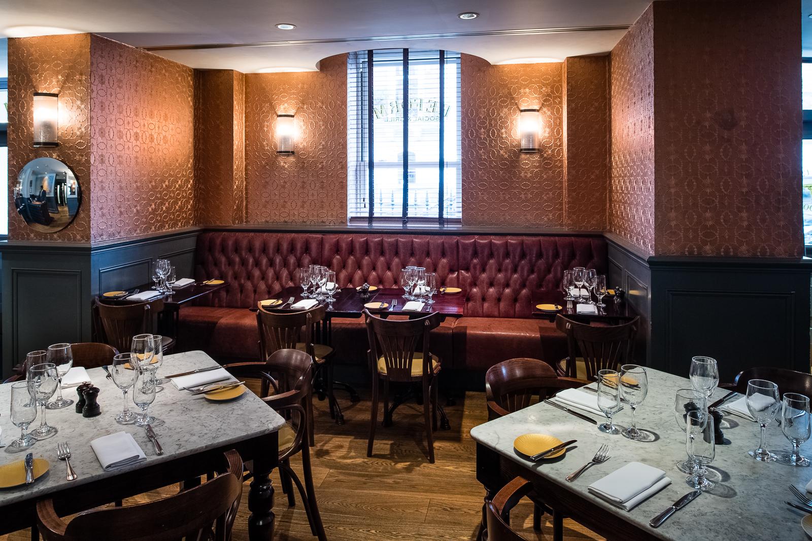 A restaurant with tufted red leather banquette seating, marble tables, and patterned wallpaper at The Mandeville Hotel.