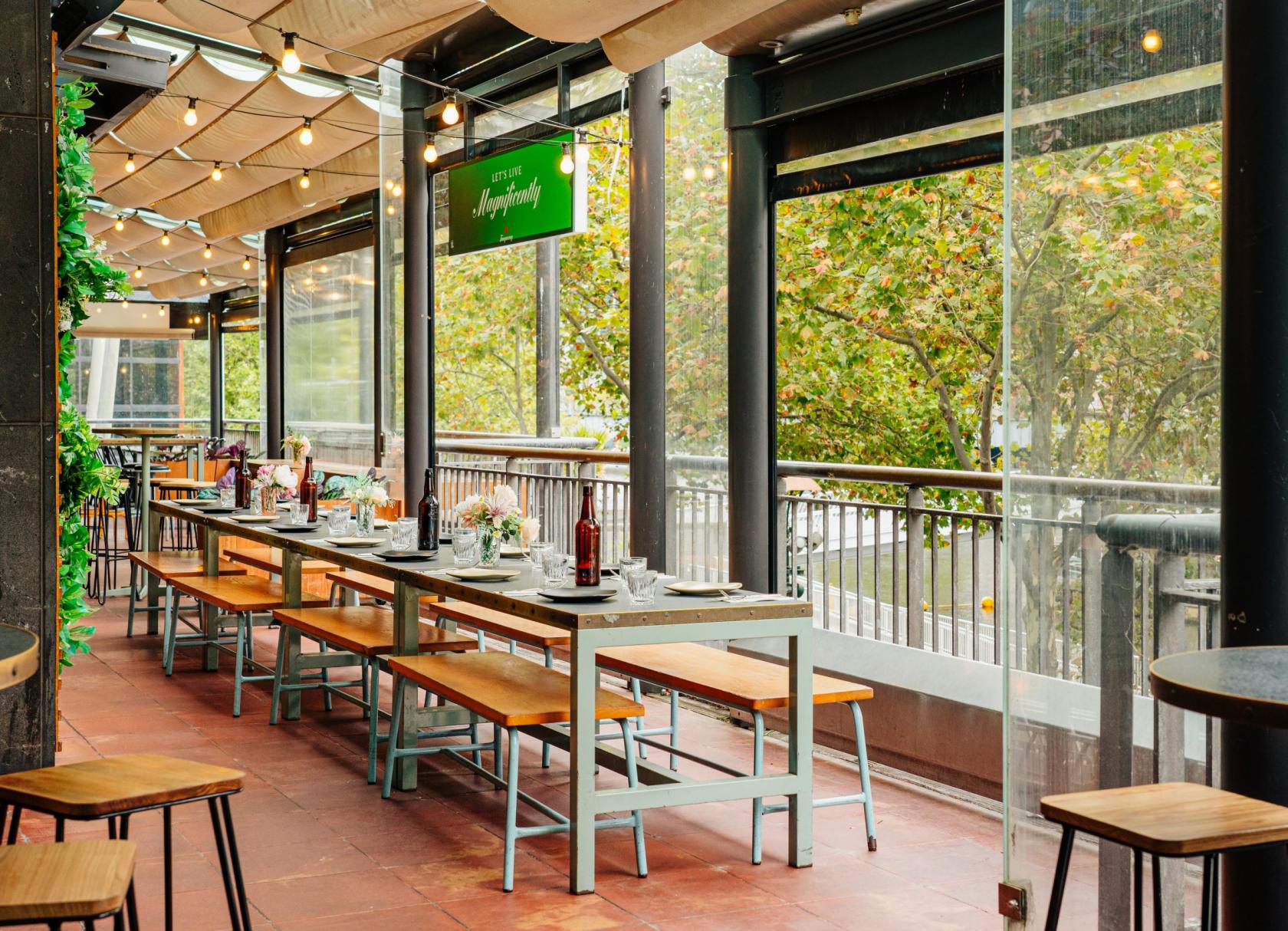 A restaurant terrace with long wooden tables, bench seating, string lights, and glass panels at Hophaus.