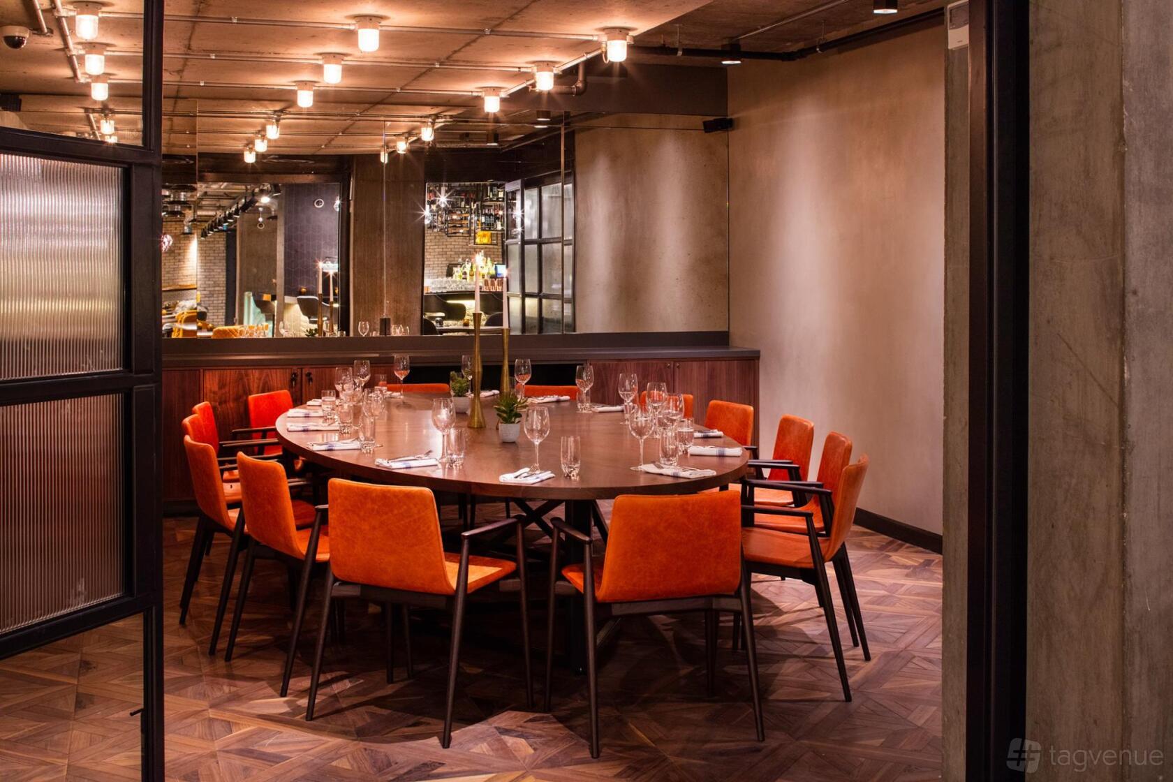A dining room with a round wooden table, orange velvet chairs, and overhead track lighting at Drake & Morgan at King's Cross.