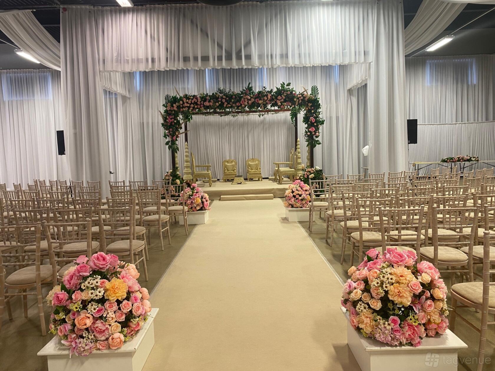 A hall with white draped walls, floral aisle arrangements, and a flower-adorned stage arch at Leicester Racecourse The Kube.
