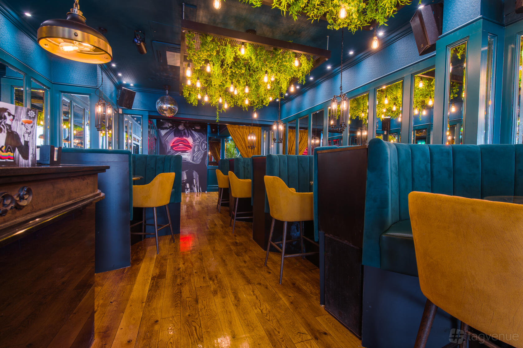 A bar lounge with teal booth seating, yellow chairs, mirrored walls, and hanging greenery at Revolution Brighton.