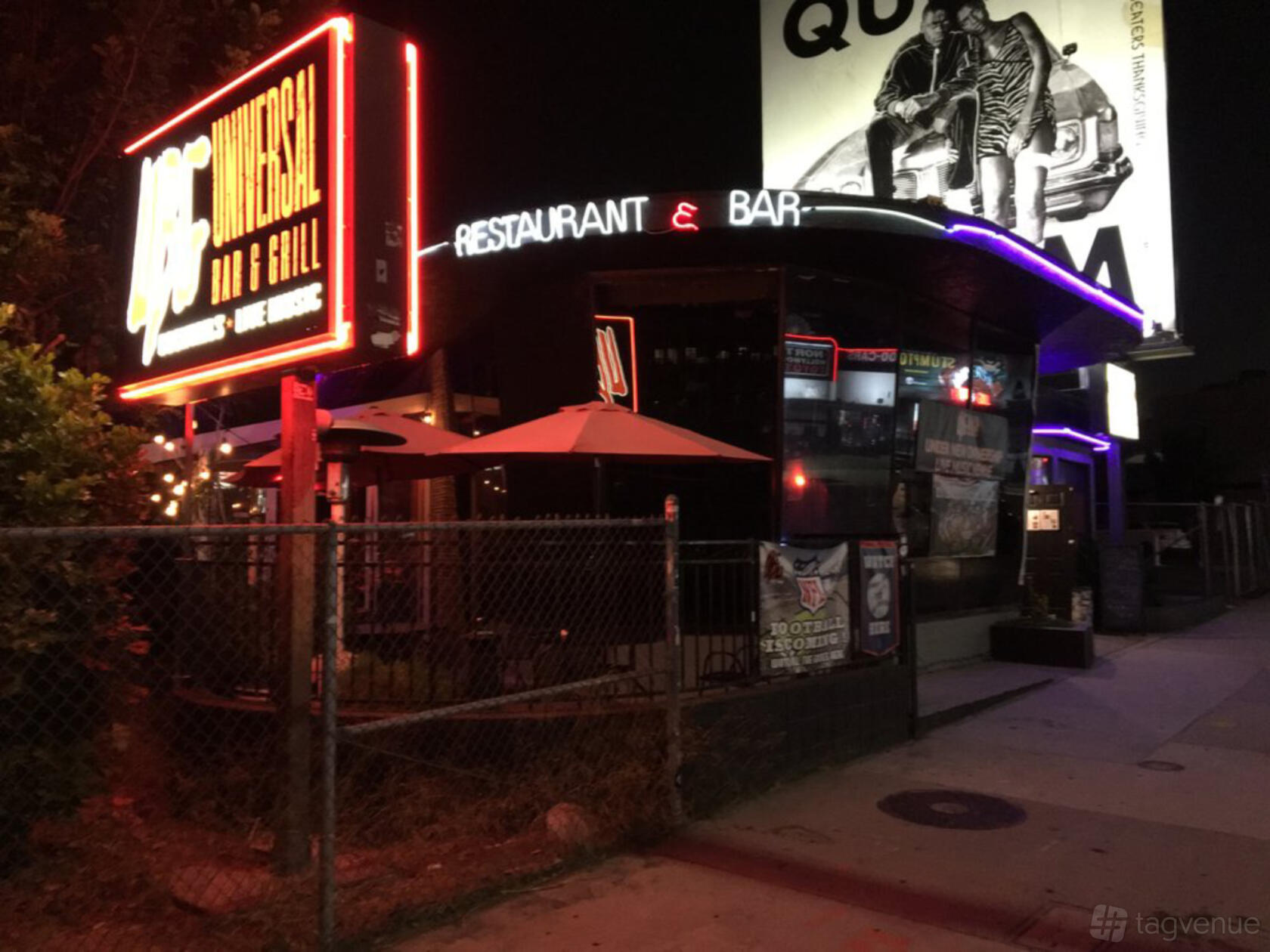 An exterior of a pub with neon signage, umbrellas on the patio, and a sidewalk entrance at Universal Bar and Grill.