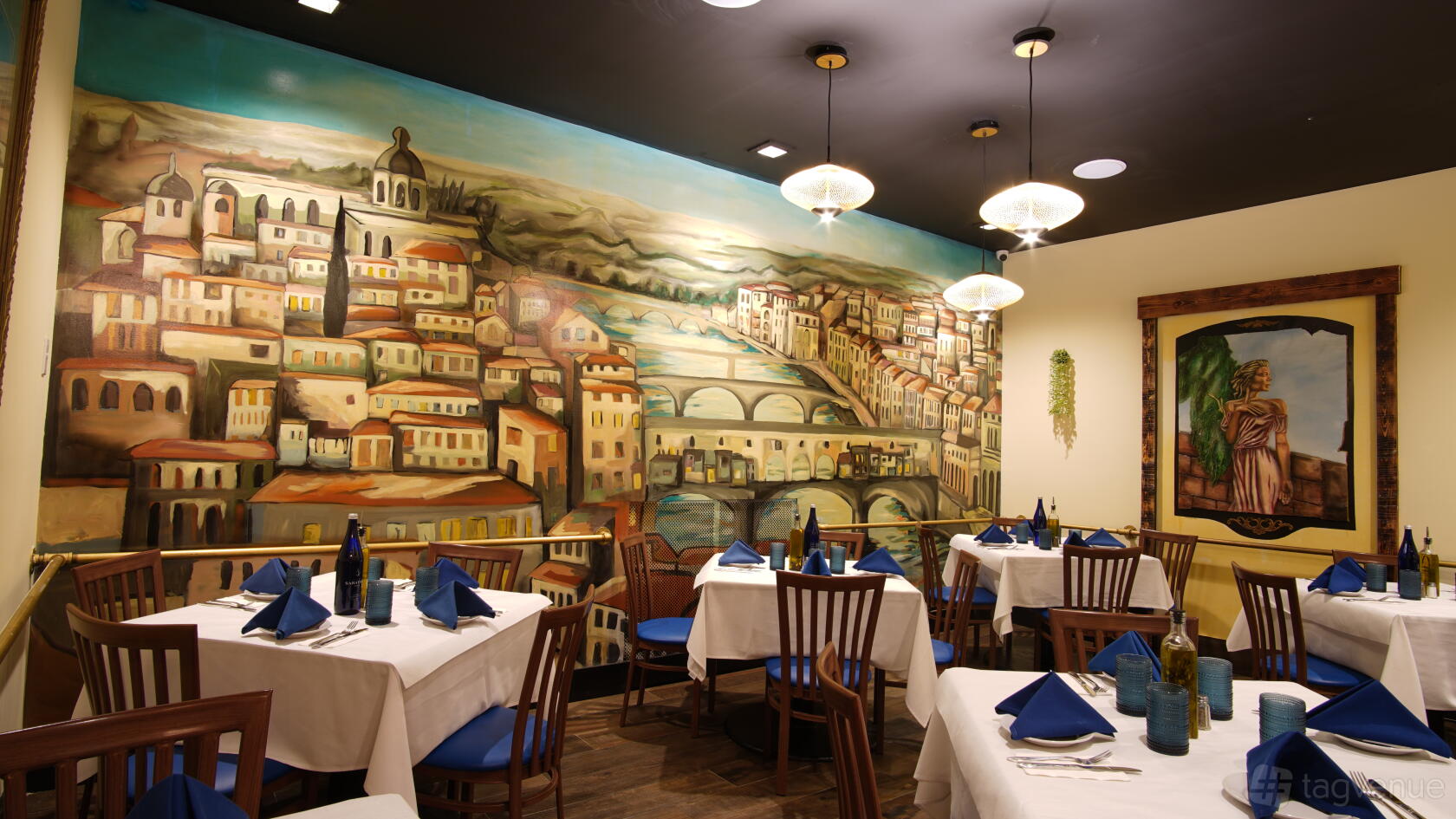 A restaurant dining room with a large Italian city mural, wooden chairs, and tables set with blue napkins at Casa D' Angelo.