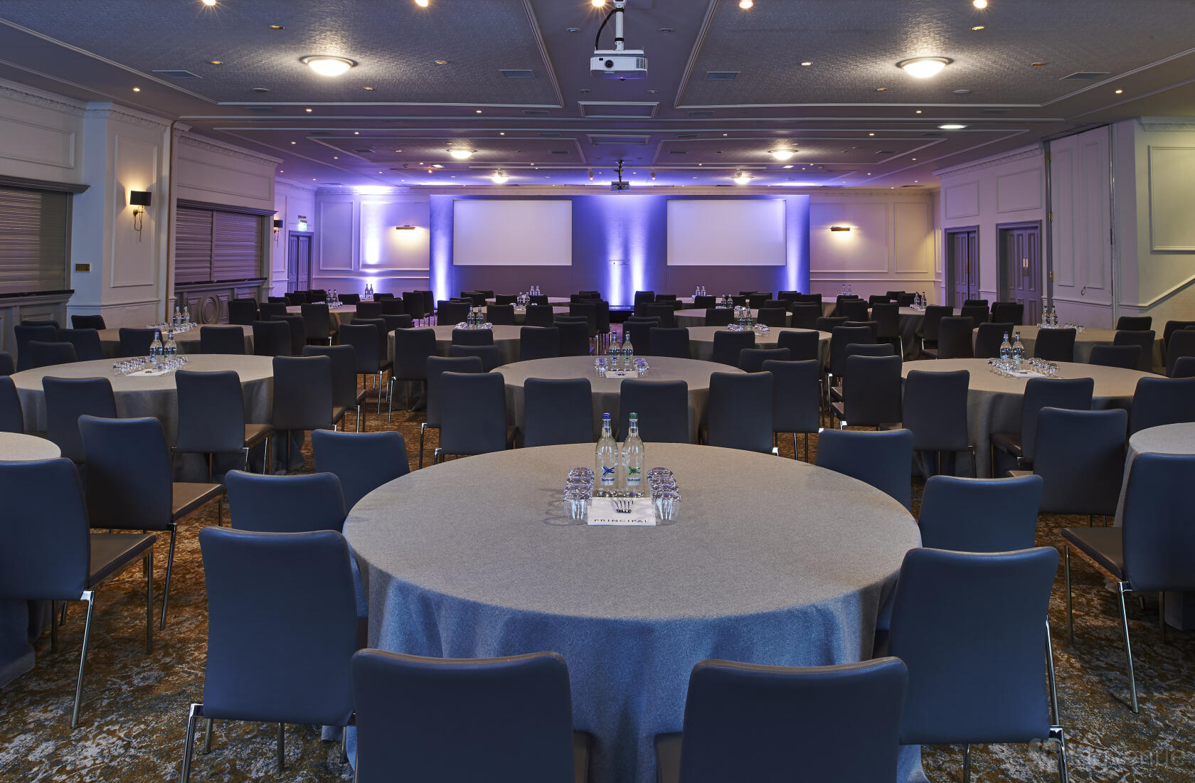 A hotel hall with round tables covered in grey linens, water bottles, and projection screens at Kimpton Charlotte Square Edinburgh.