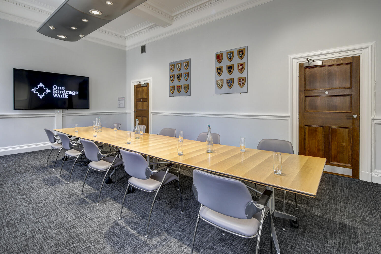 A meeting room with a long wooden table, grey chairs, wall-mounted shields, and a large screen at One Birdcage Walk.
