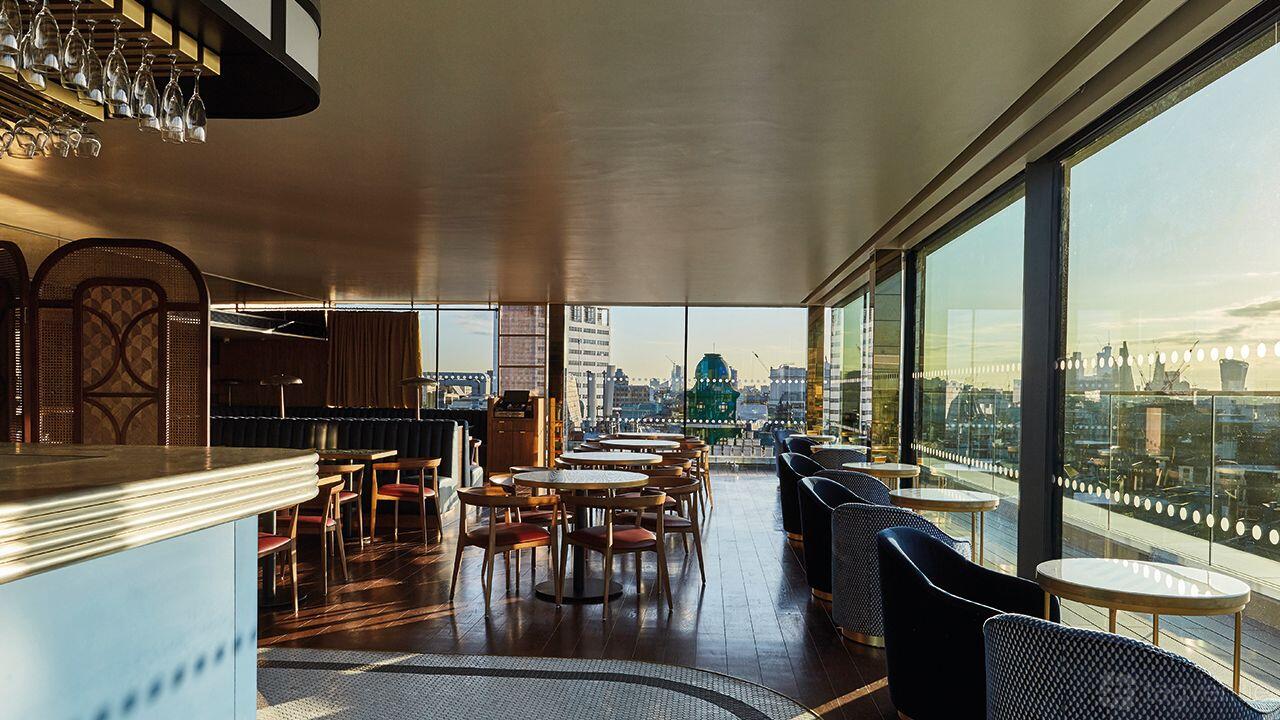 A rooftop cocktail bar with floor-to-ceiling windows, round wooden tables, and city skyline views at LSQRooftop.