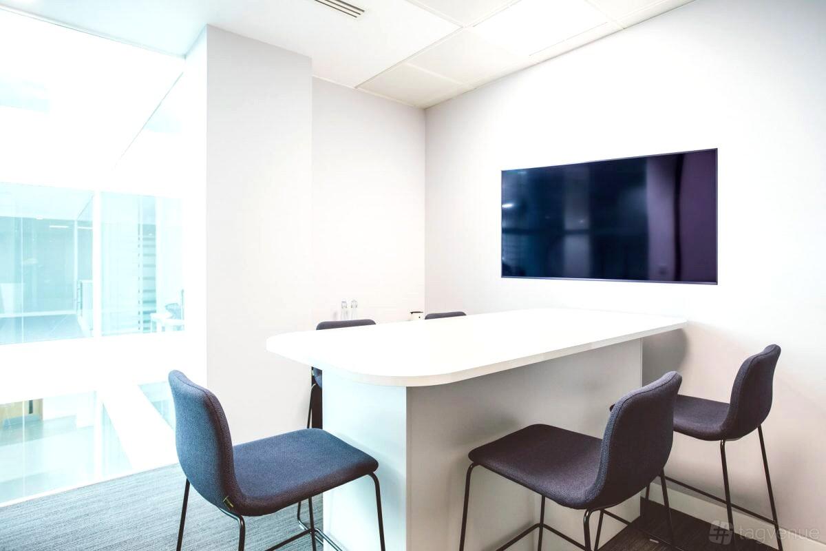 A meeting room with a high white table, four dark chairs, and a wall-mounted flatscreen at Orega Uxbridge.