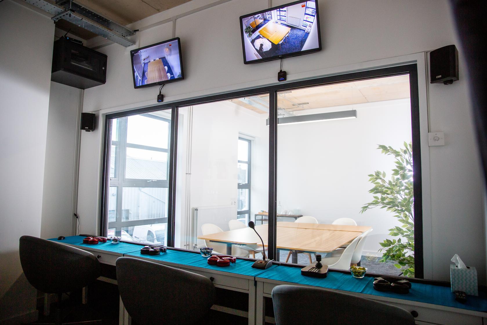 A meeting room with a glass observation window, wall-mounted screens, and a table with chairs at Human Factors Lab and Conference Centre.