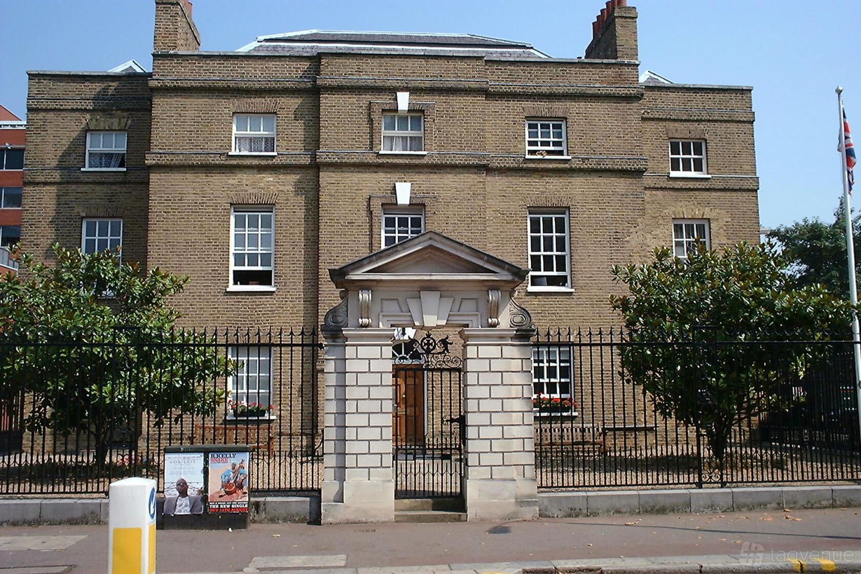 A historic brick building with white-trimmed windows, black iron fencing, and a flagpole at Fulham, Fulham High Street.