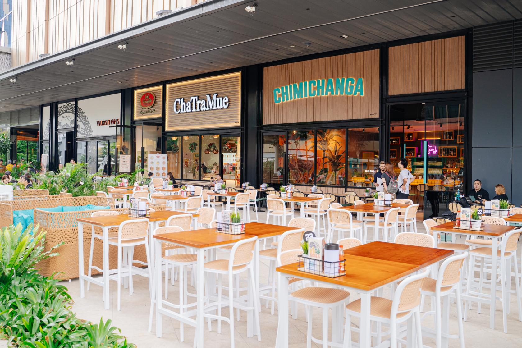 An outdoor restaurant seating area with wooden tables, white chairs, and greenery at Chimichanga Paya Lebar Quarters.
