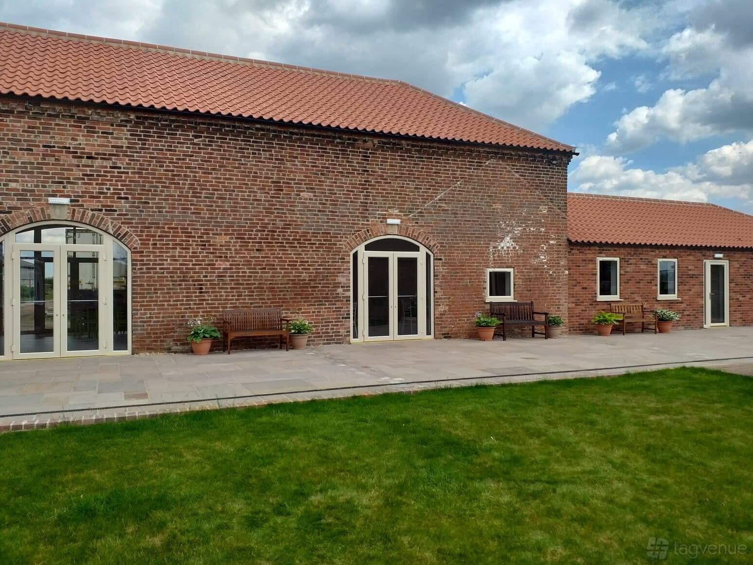 A red brick barn with arched doors, patio benches, potted plants, and a tiled walkway at Dunstall Barn.