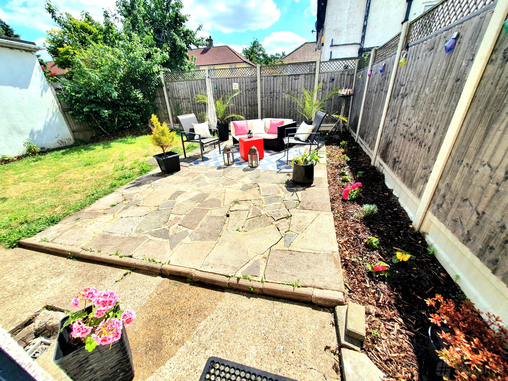 A house garden with a stone patio, outdoor seating, potted plants, and a wooden fence at 3 BED HOUSE.