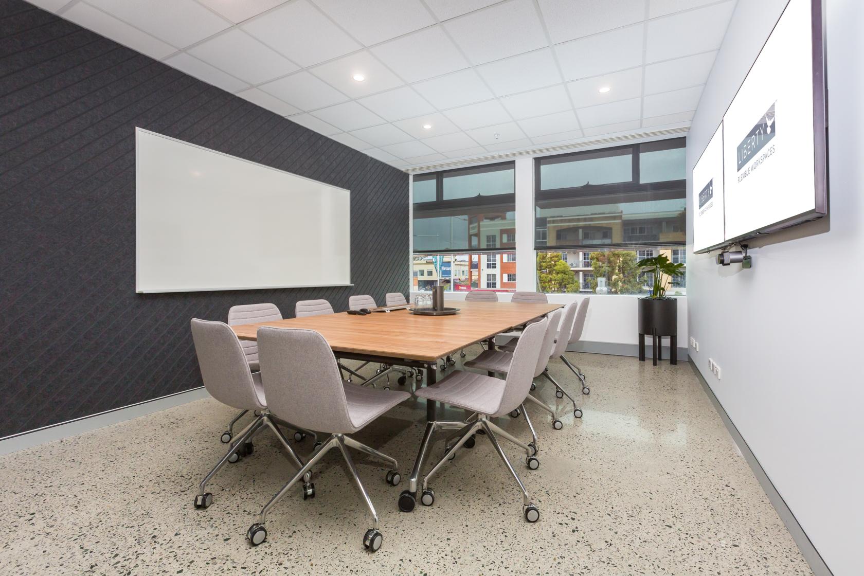 A boardroom with a large wooden meeting table, gray chairs on casters, whiteboard, and wall-mounted screen at Liberty Joondalup.